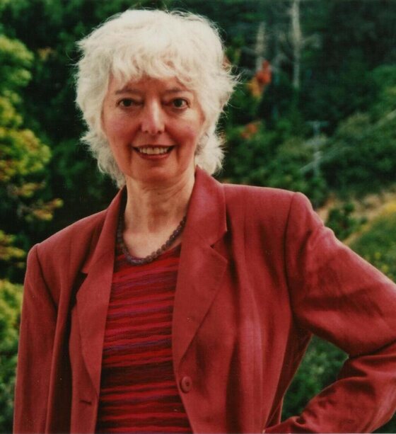 Emily Wortis Leider outdoors in a red jacket, smiling, with wooded hills behind her